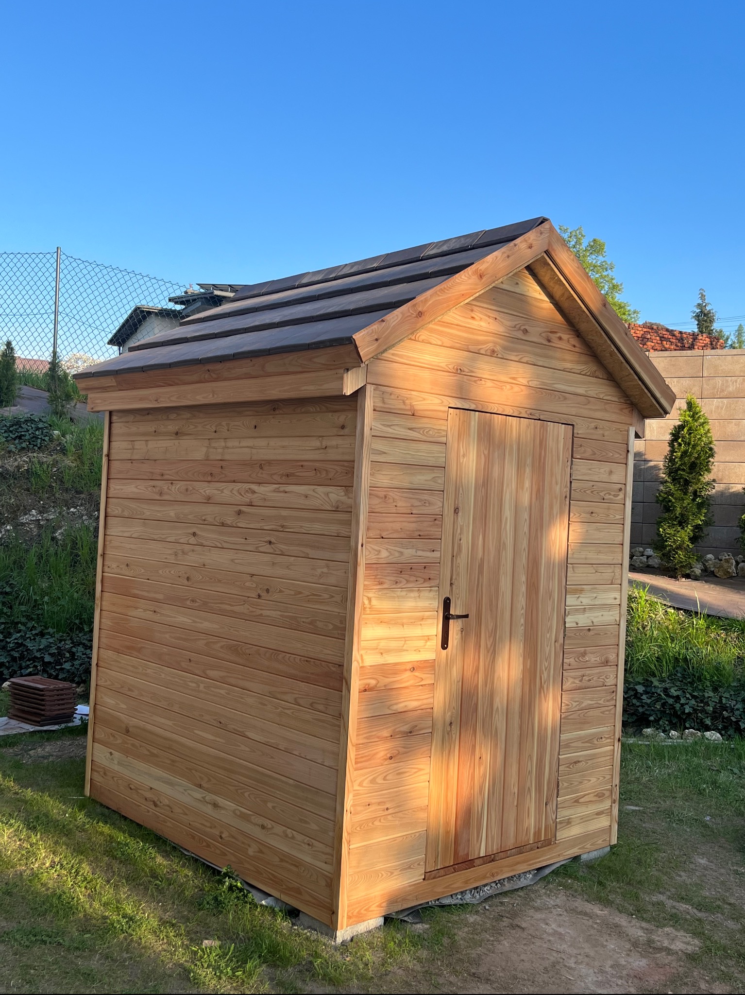 Drewniana szopa narzędziowa z brązowym dachem, umieszczona na trawniku w ogrodzie, widoczna w słonecznym świetle poranka.