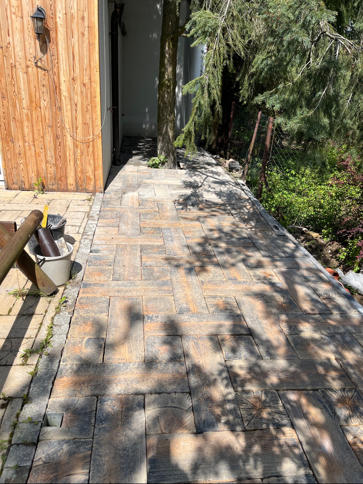 Ułożona ścieżka z kostki brukowej imitującej drewno, prowadząca obok drewnianej ściany i drzewa, z widocznymi narzędziami budowlanymi w tle.