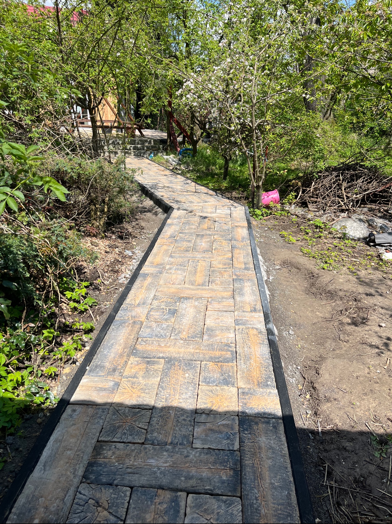 Ścieżka z kostki o nieregularnym wzorze imitującym drewno, obrzeża z czarnego tworzywa, prowadząca przez zielony ogród w kierunku drewnianej altanki.