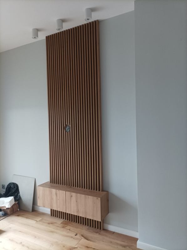 Drewniana ścianka działowa z pionowych listew zamontowana na ścianie, z szafką RTV u dołu i otworem na kable pośrodku, w nowoczesnym wnętrzu.