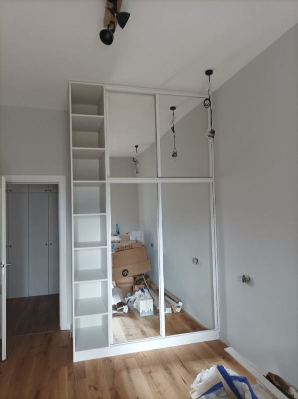 Biała szafa wnękowa z lustrzanymi drzwiami przesuwnymi w trakcie montażu, widoczne kartony i narzędzia na podłodze, drewniana podłoga.