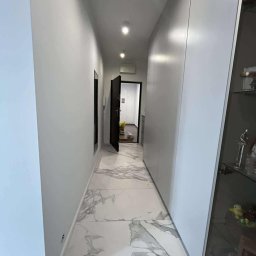 Bartłomiej Biały - Nowoczesny korytarz z białymi ścianami, marmurową podłogą i zabudowaną szafą. Otwarta czarna rama drzwi prowadzi do jasnego pomieszczenia w głębi.