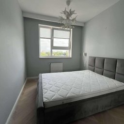 Bartłomiej Biały - Pokój po remoncie: szare ściany, drewniana podłoga w jodełkę, łóżko z szarym zagłówkiem, okno z żaluzjami i designerska lampa sufitowa.