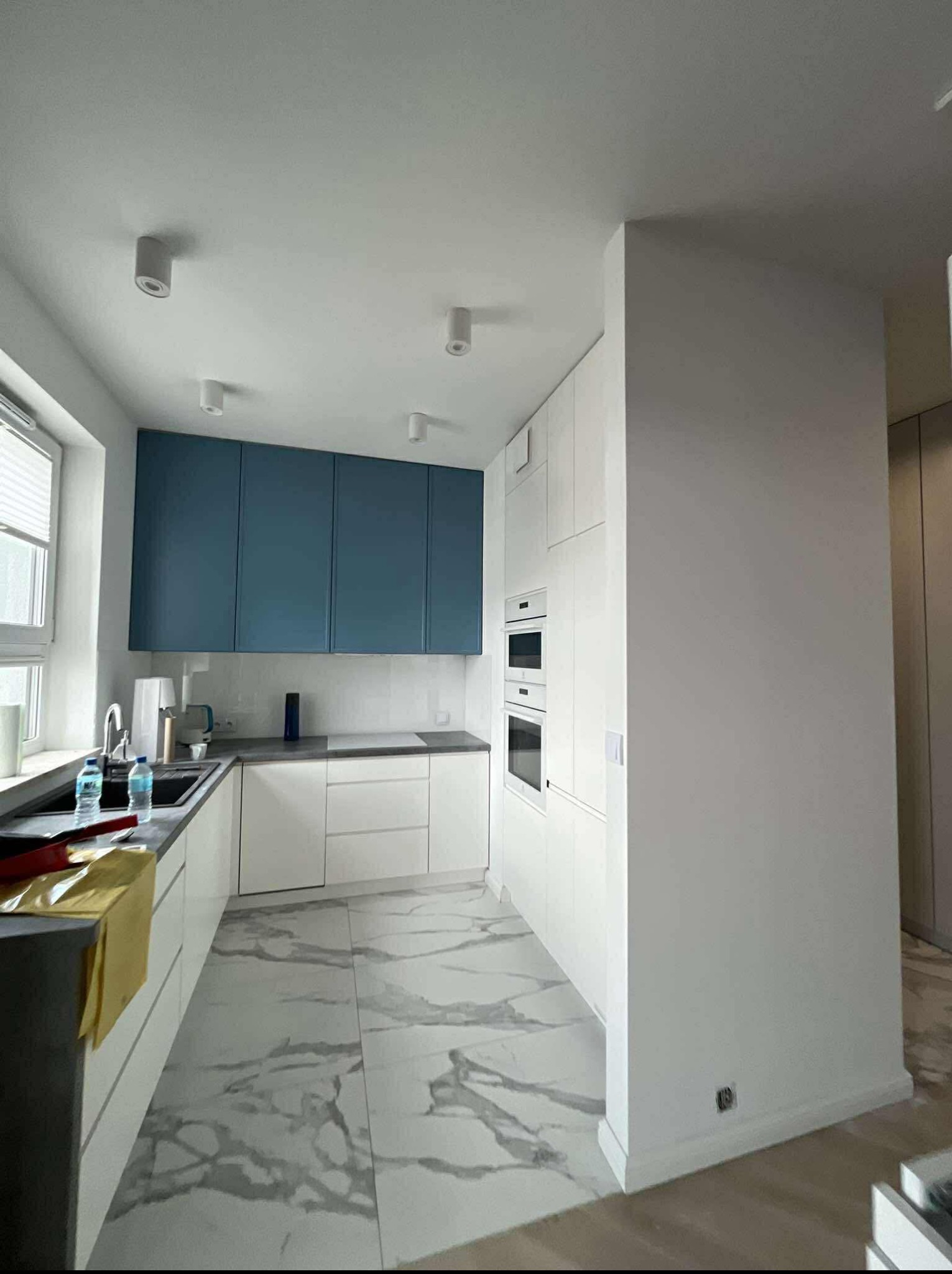 Nowoczesna kuchnia z niebieskimi szafkami i marmurową podłogą w Ostrowie Lubelskim. Białe fronty dolnych szafek i zabudowane sprzęty AGD.