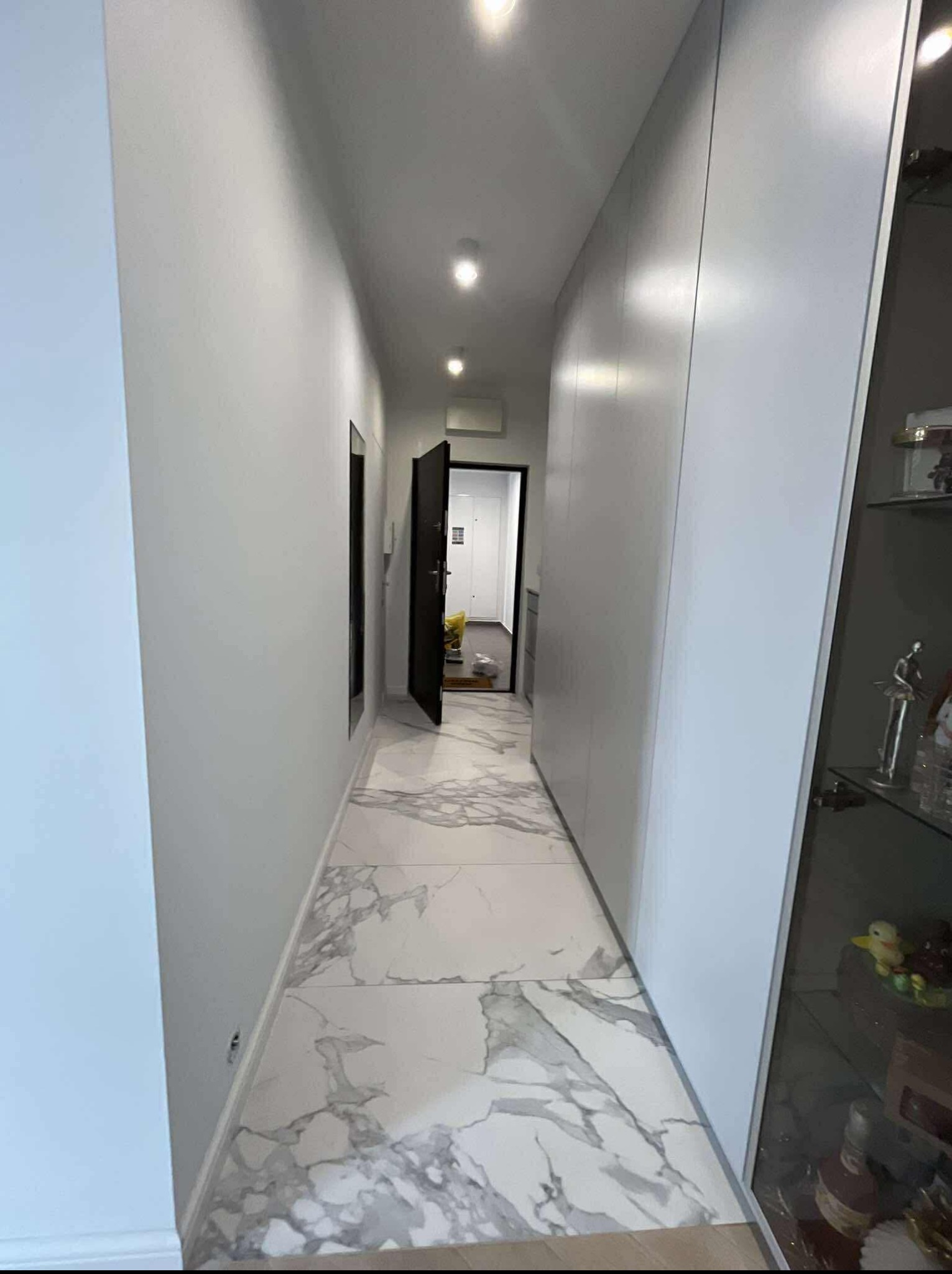 Nowoczesny korytarz z białymi ścianami, marmurową podłogą i zabudowaną szafą. Otwarta czarna rama drzwi prowadzi do jasnego pomieszczenia w głębi.