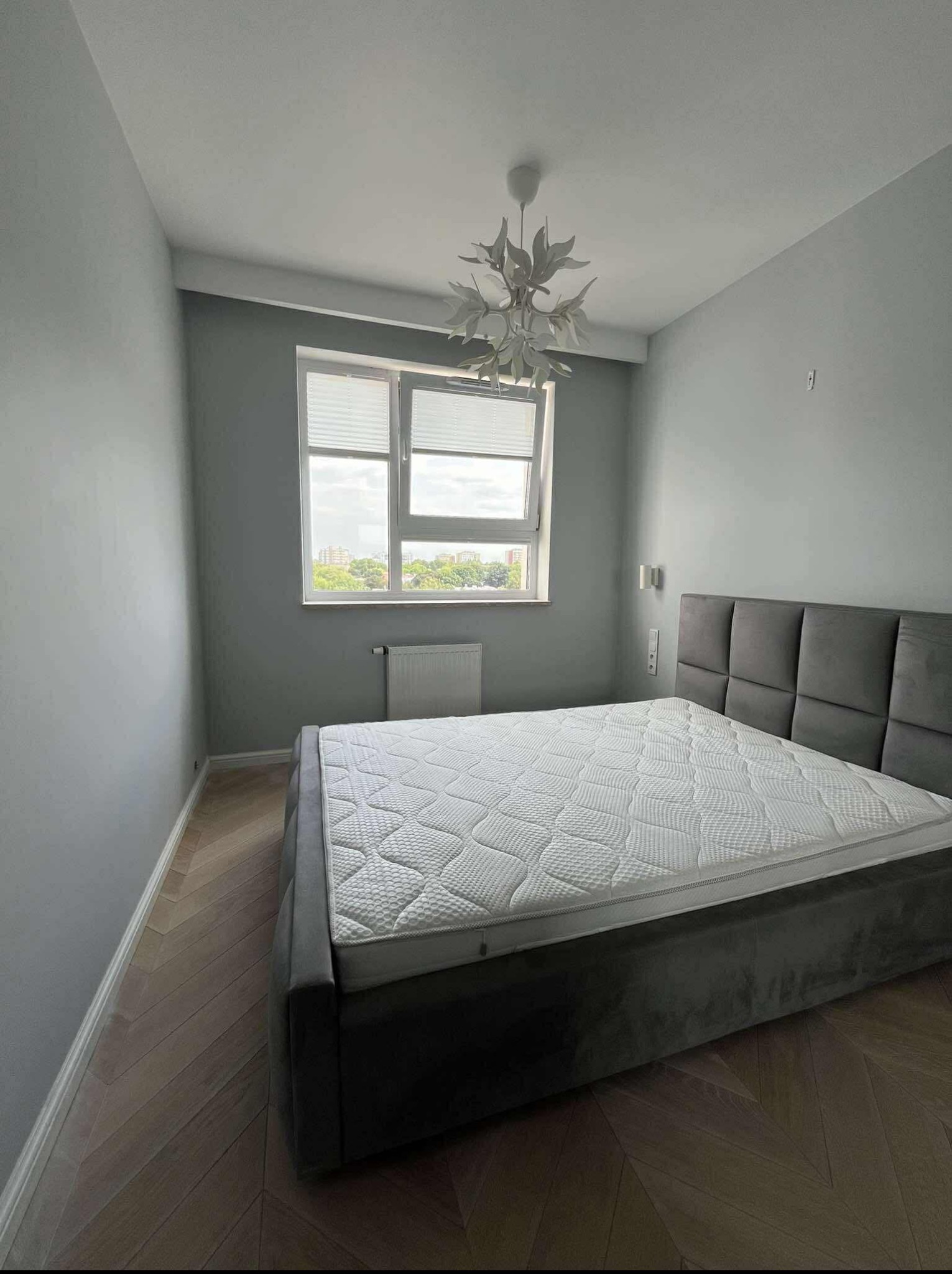 Pokój po remoncie: szare ściany, drewniana podłoga w jodełkę, łóżko z szarym zagłówkiem, okno z żaluzjami i designerska lampa sufitowa.