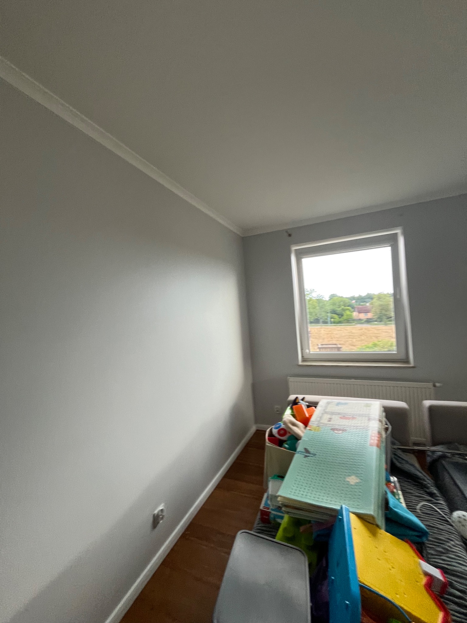 Jasnoszara ściana po malowaniu z widoczną listwą przypodłogową i oknem z widokiem na zewnątrz. W pokoju panuje lekki nieład, co sugeruje, że jest to pokój dziecięcy.