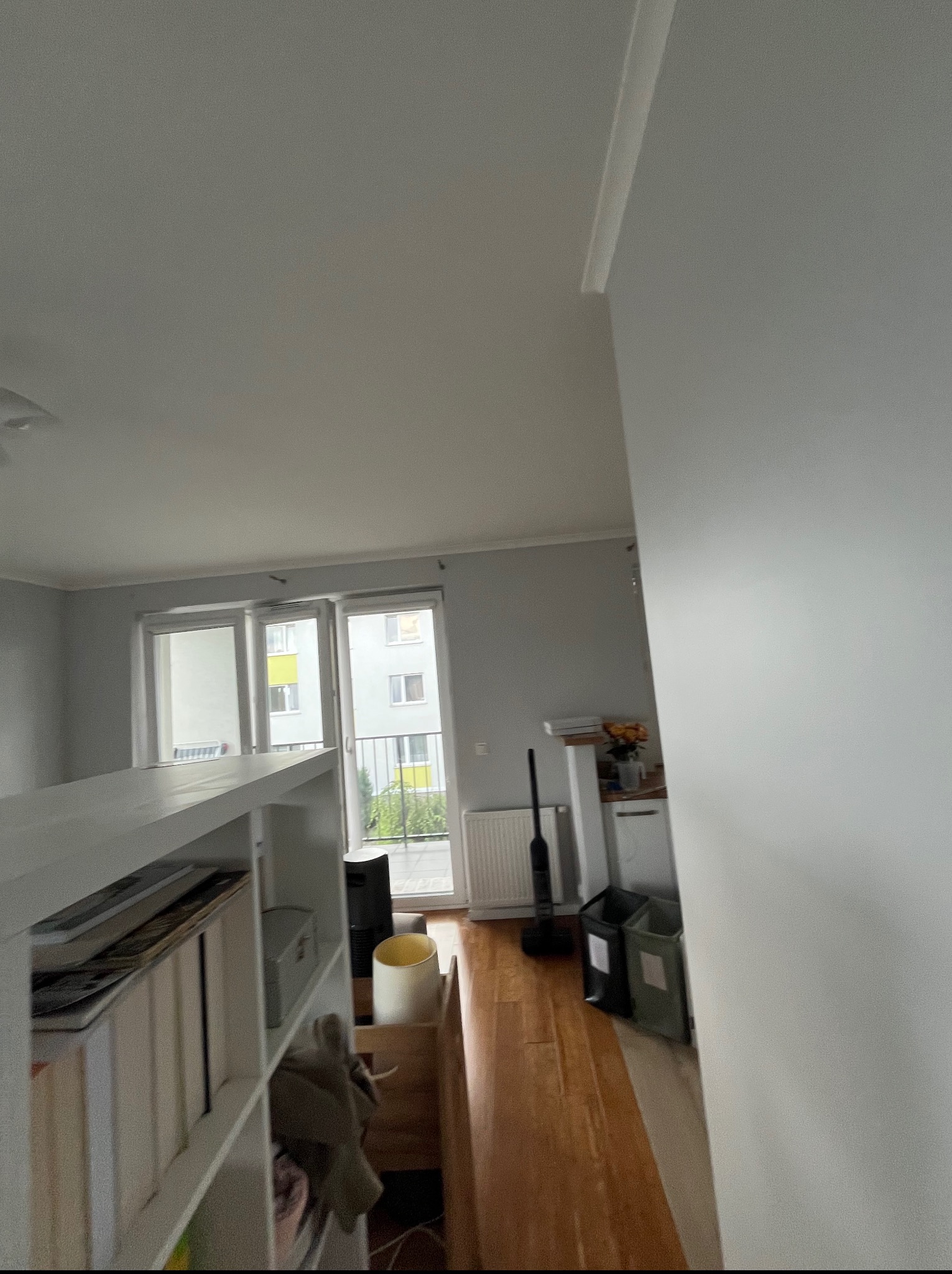 Jasne wnętrze po odświeżeniu: widok na balkon z drewnianą podłogą, białe ściany i regał z książkami. Minimalistyczny design, świeżo po pracach wykończeniowych.