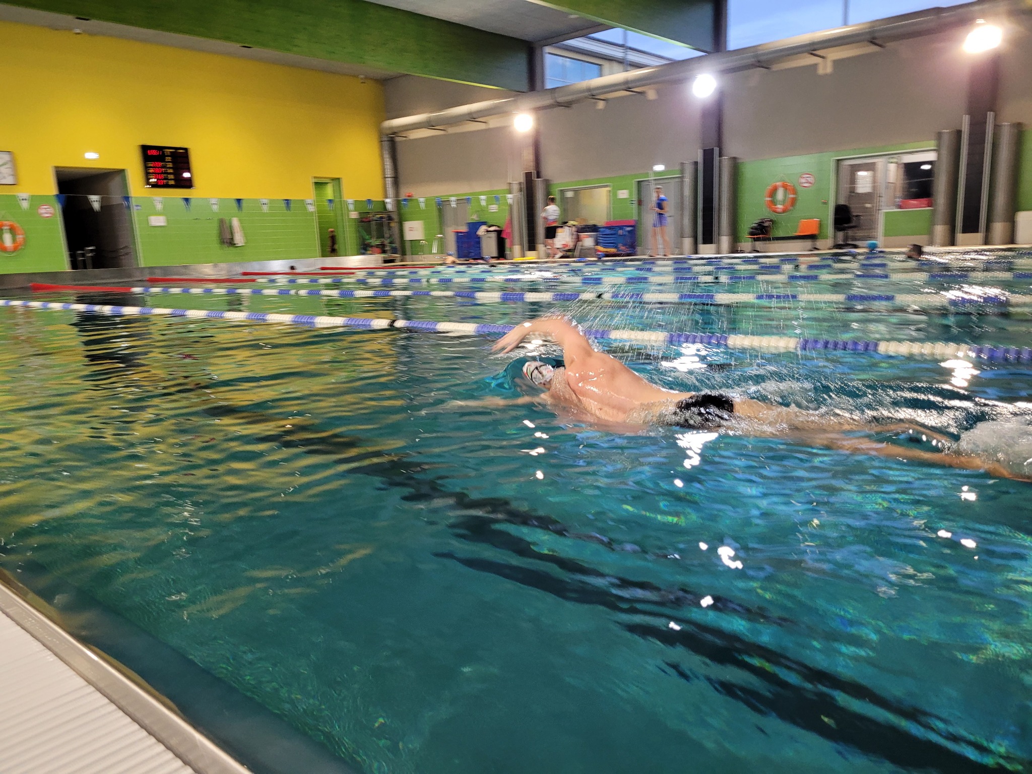 Pływak w trakcie treningu stylem dowolnym w krytej pływalni, widoczne tory pływackie i elektroniczny zegar wyników na ścianie.