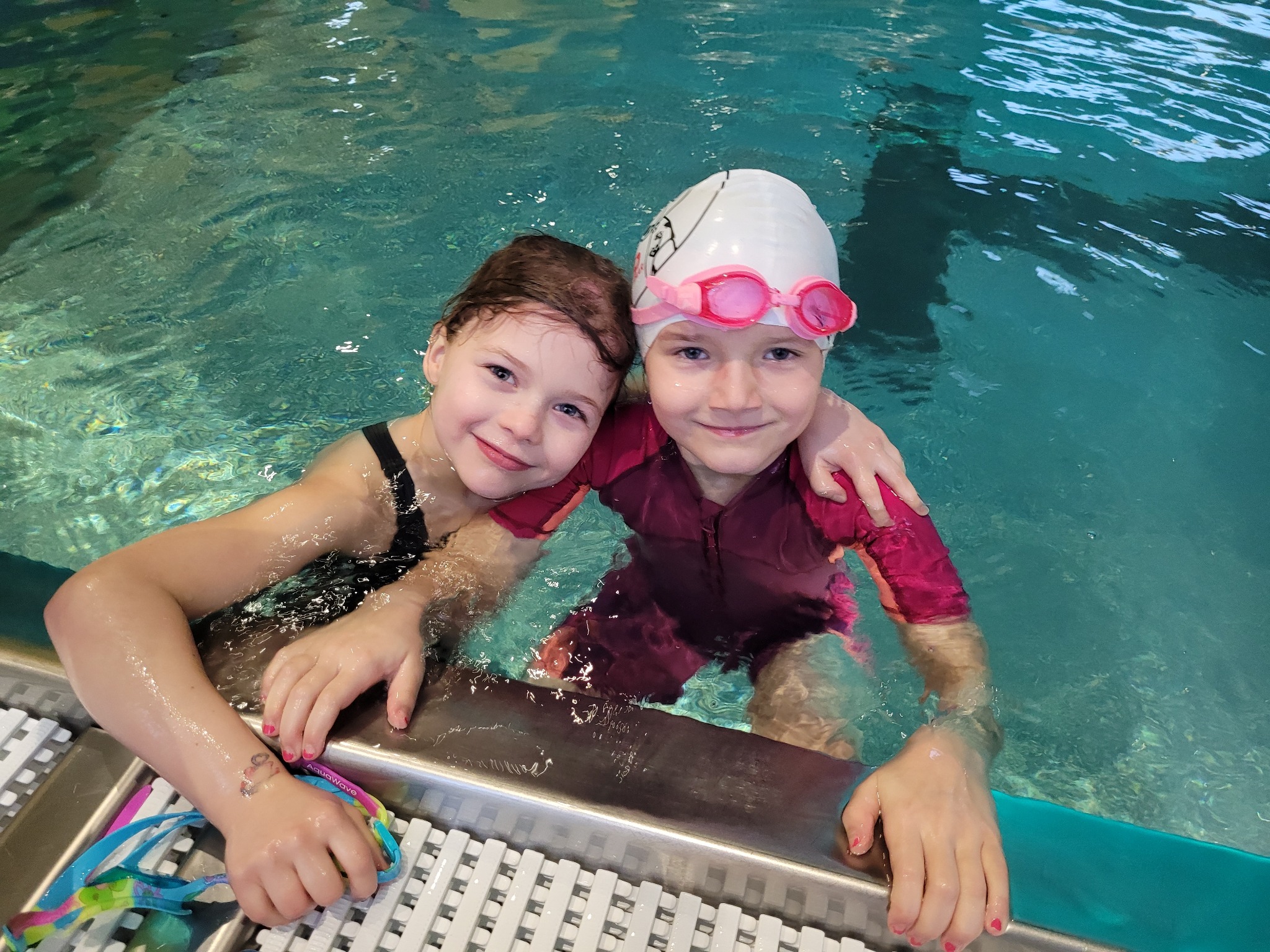 Dwie uśmiechnięte dziewczynki w basenie, jedna w czarnym stroju kąpielowym, druga w różowym z czepkiem i różowymi okularami, opierają się o krawędź basenu.