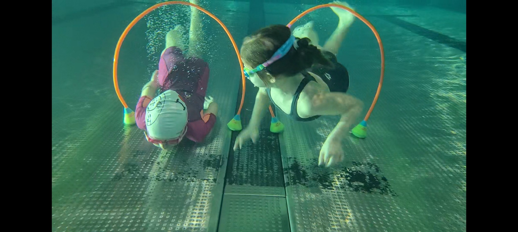 Dwie dziewczynki w czepkach i strojach kąpielowych nurkują pod wodą, przechodząc przez pomarańczowe obręcze z zielonymi obciążnikami, widoczne bąbelki powietrza.
