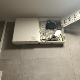 WOL-TECH Julian Wolski - Otwarta skrzynka instalacyjna na ścianie z routerem, zasilaczami i przewodami wewnątrz. Widoczna biała obudowa i szare płytki podłogowe.