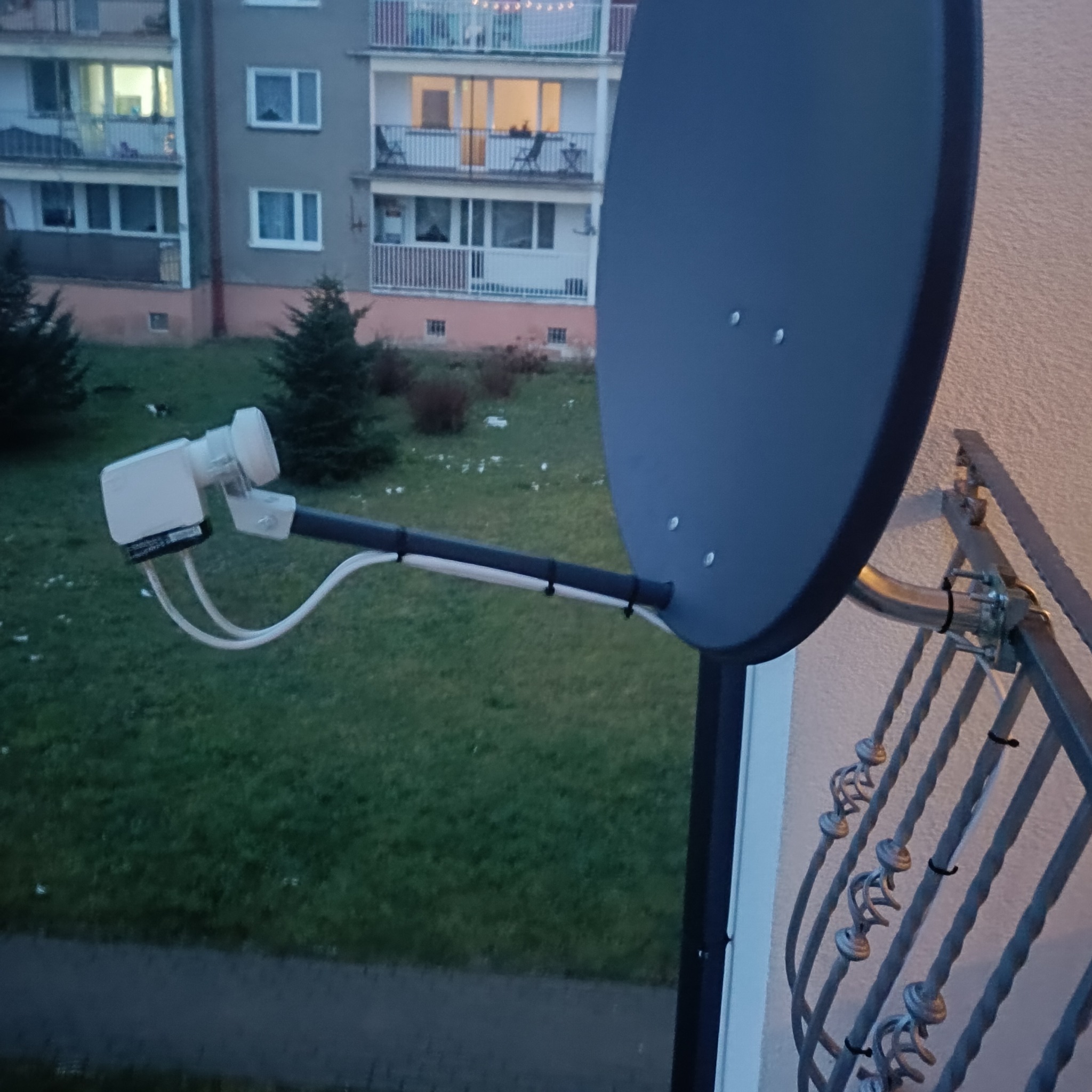Antena satelitarna zamontowana na balkonie z widokiem na blok mieszkalny i zieleń.