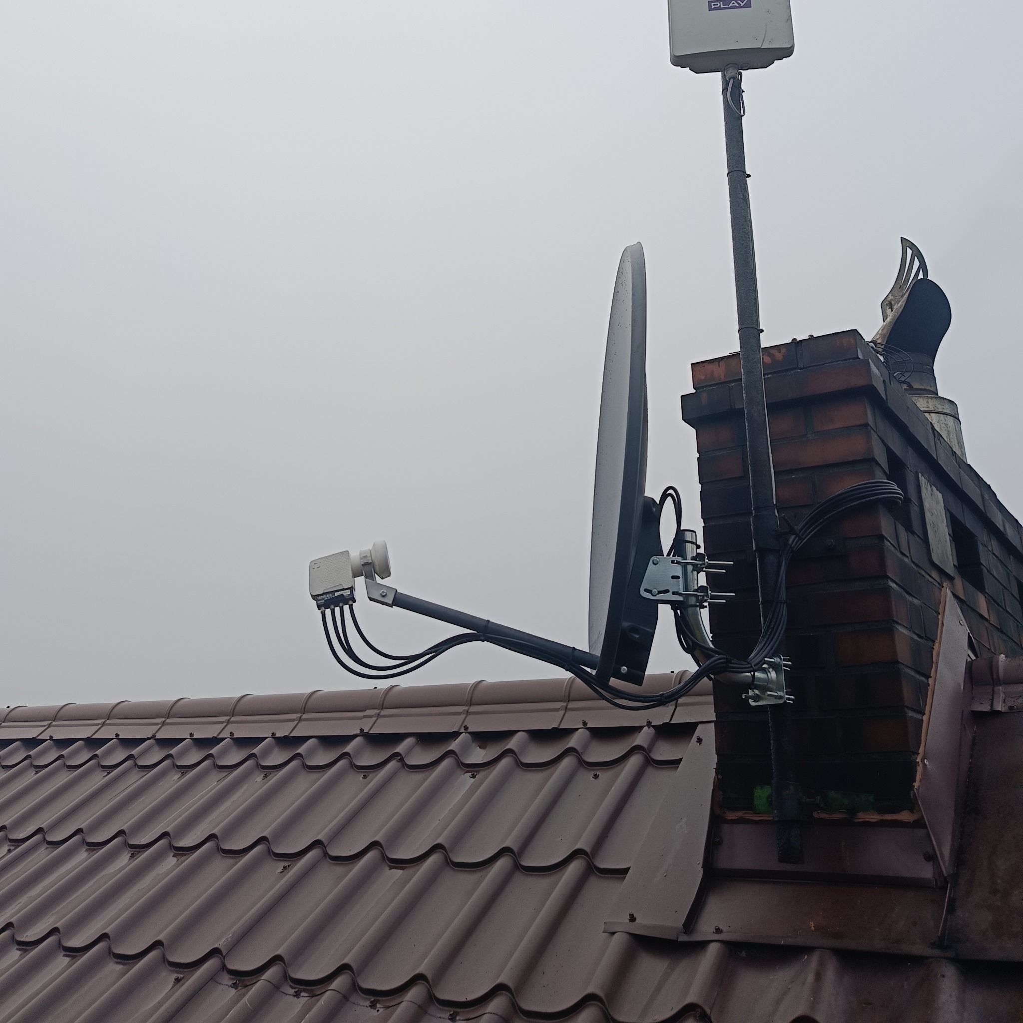 Antena satelitarna i antena LTE Play zamontowane na kominie obok brązowego dachu z blachodachówki w pochmurny dzień.