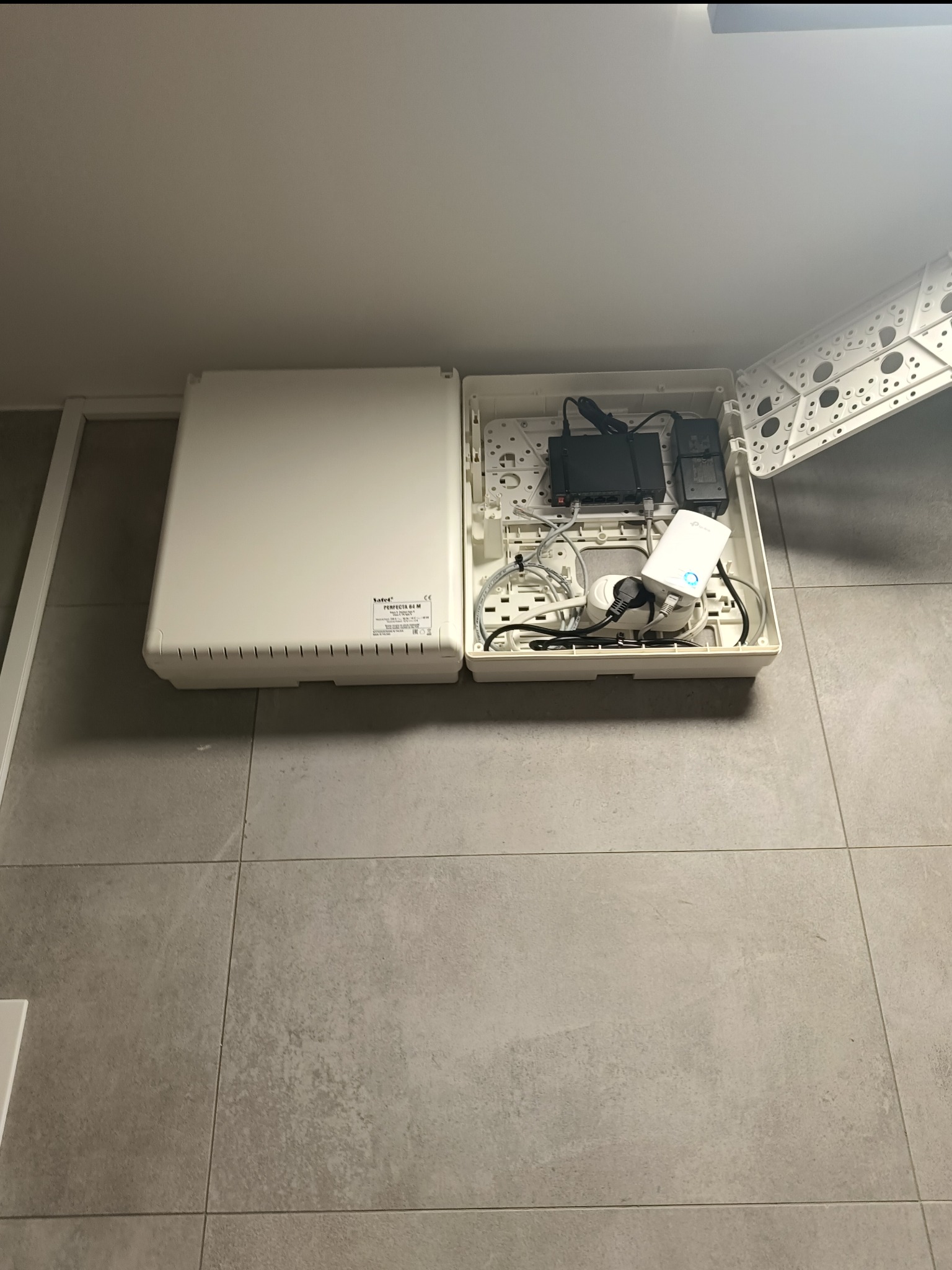 Otwarta skrzynka instalacyjna na ścianie z routerem, zasilaczami i przewodami wewnątrz. Widoczna biała obudowa i szare płytki podłogowe.