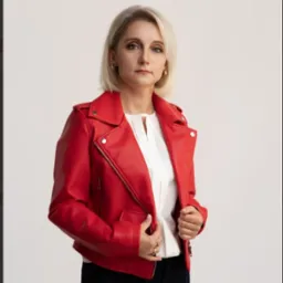 Portret kobiety w czerwonej skórzanej kurtce na jasnym tle. Uśmiechnięta, z krótkimi blond włosami i delikatnym makijażem.