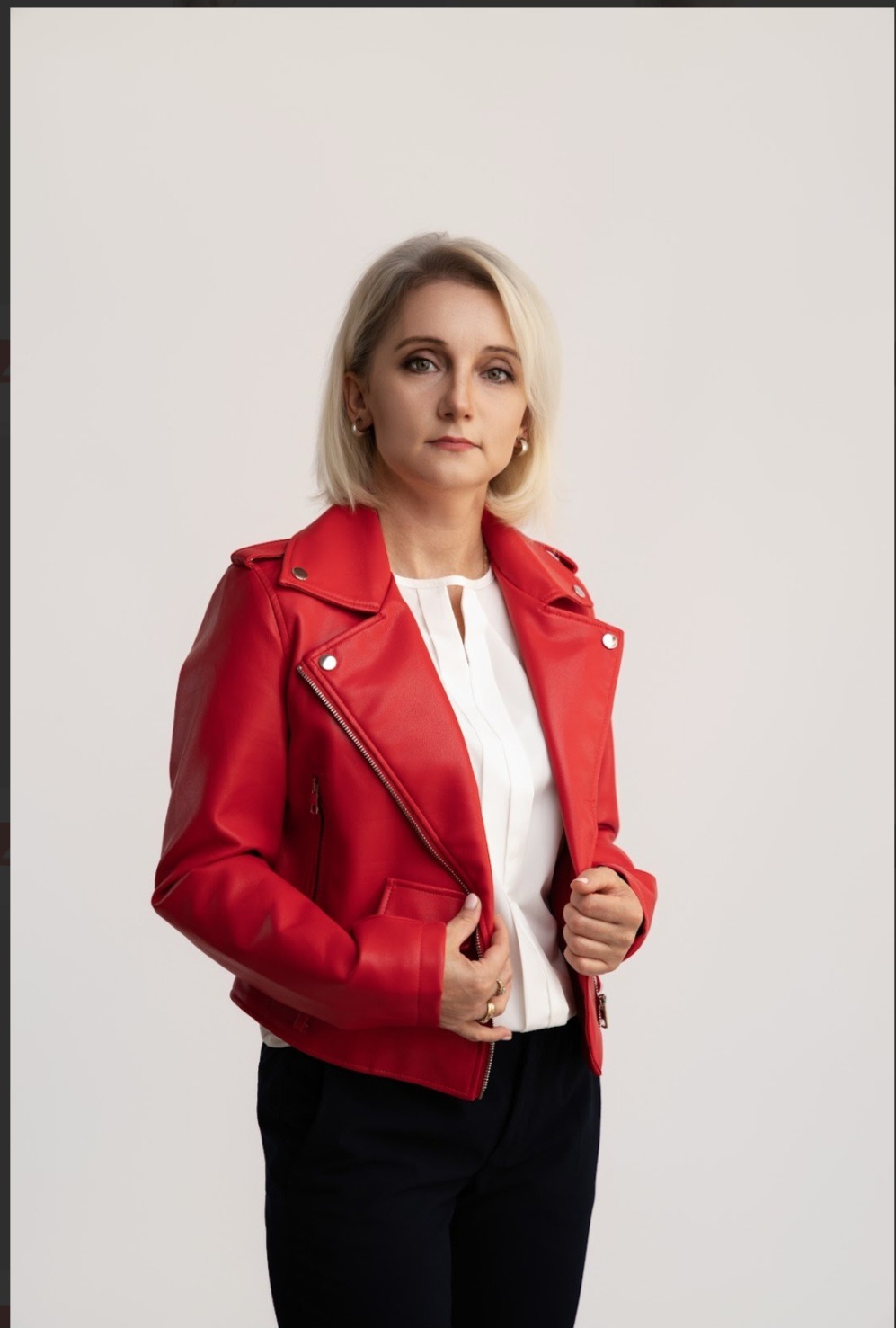 Portret kobiety w czerwonej skórzanej kurtce na jasnym tle. Uśmiechnięta, z krótkimi blond włosami i delikatnym makijażem.