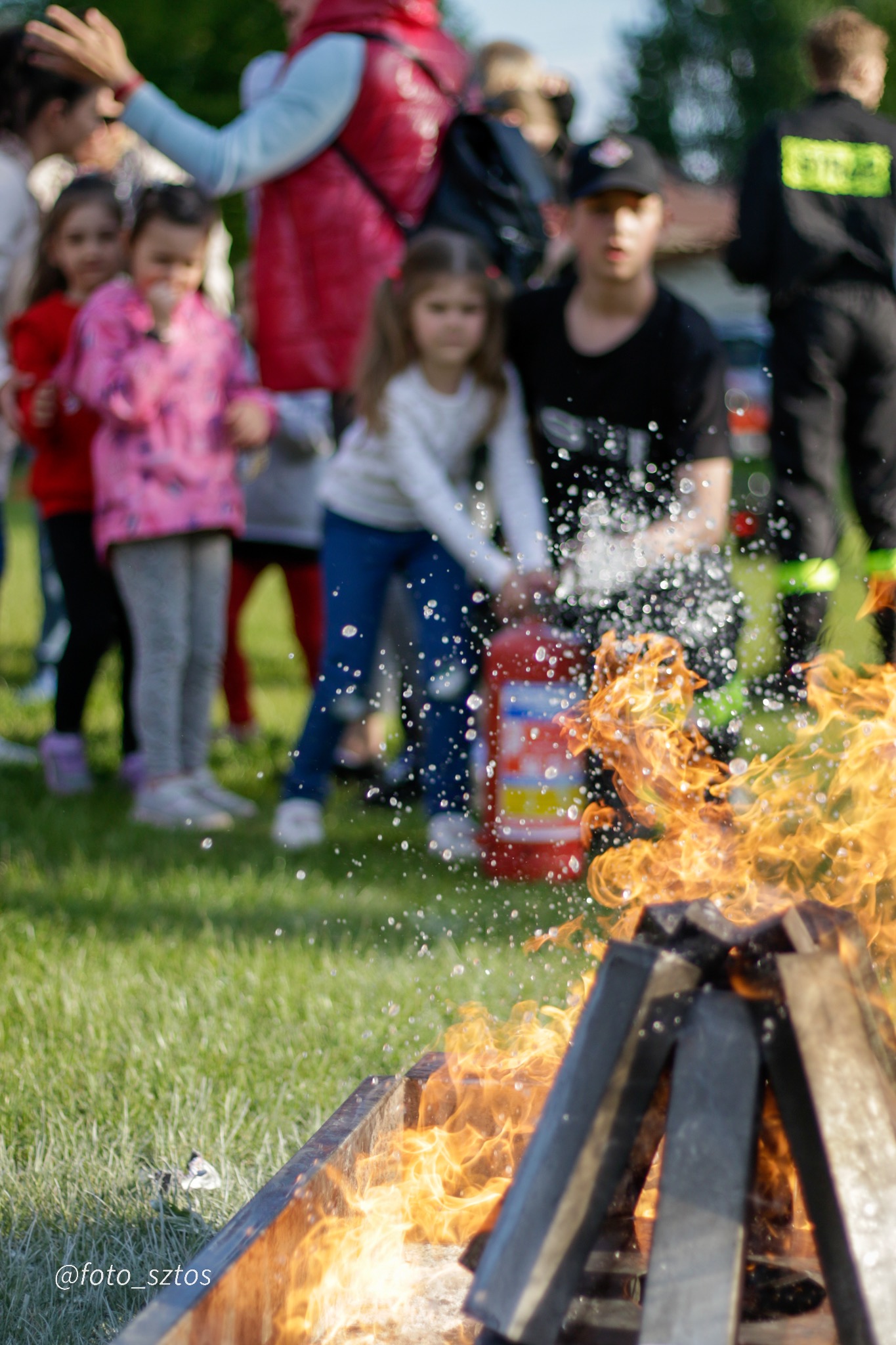Dzieci obserwują demonstrację gaszenia ognia gaśnicą, na tle zielonej trawy i innych osób.