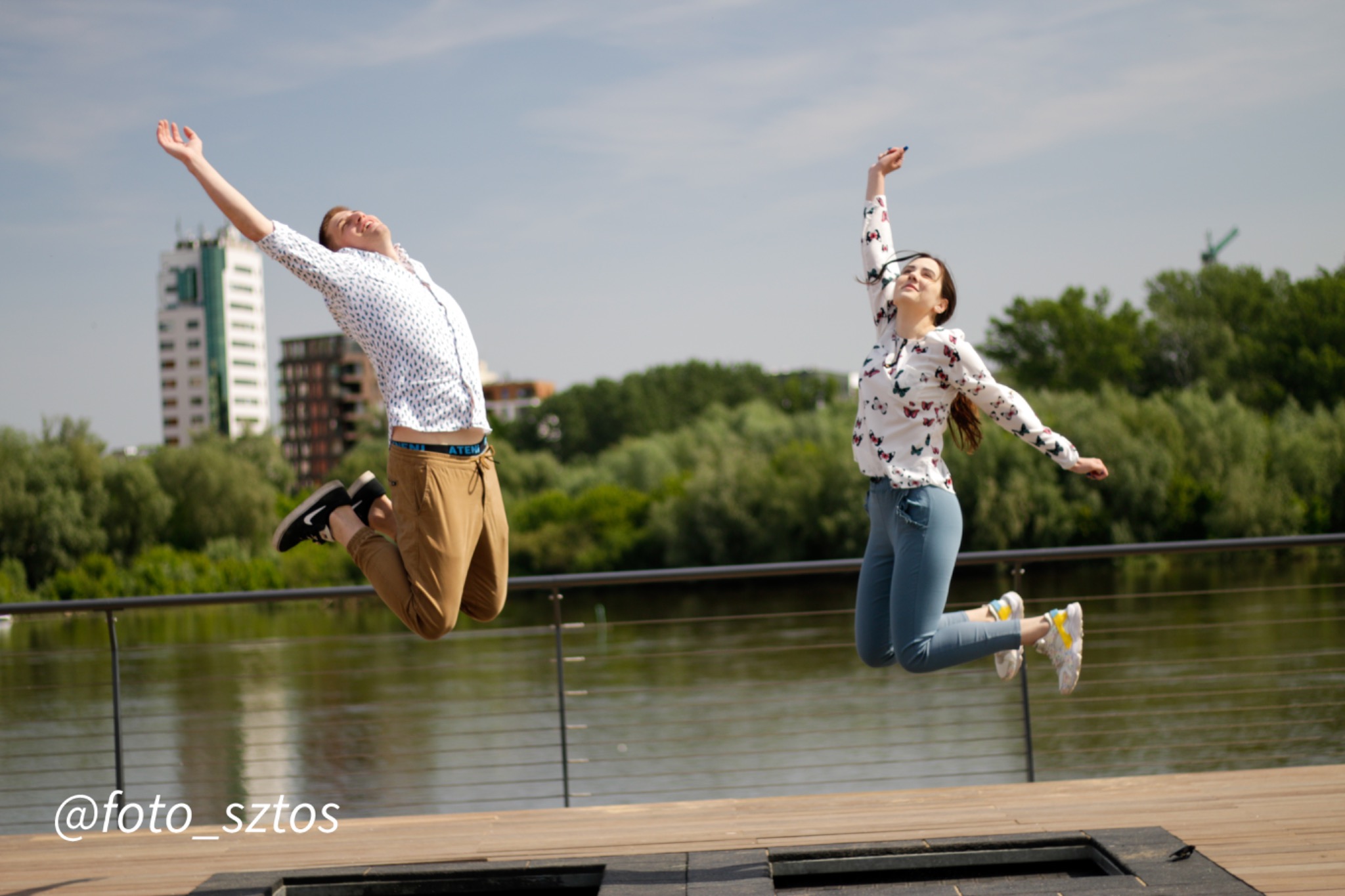 Para wesoło skacze na trampolinie nad rzeką w słoneczny dzień, w tle budynki i zieleń.
