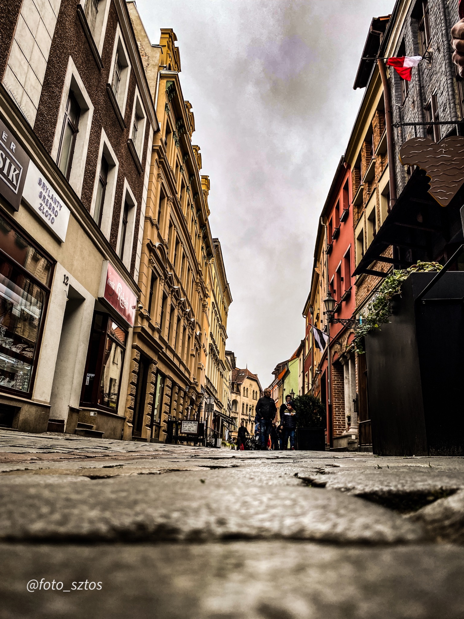 Ulica w starym mieście z perspektywy poziomu kostki brukowej, widoczne kamienice o różnej kolorystyce i architekturze, osoby spacerujące w oddali pod pochmurnym niebem.