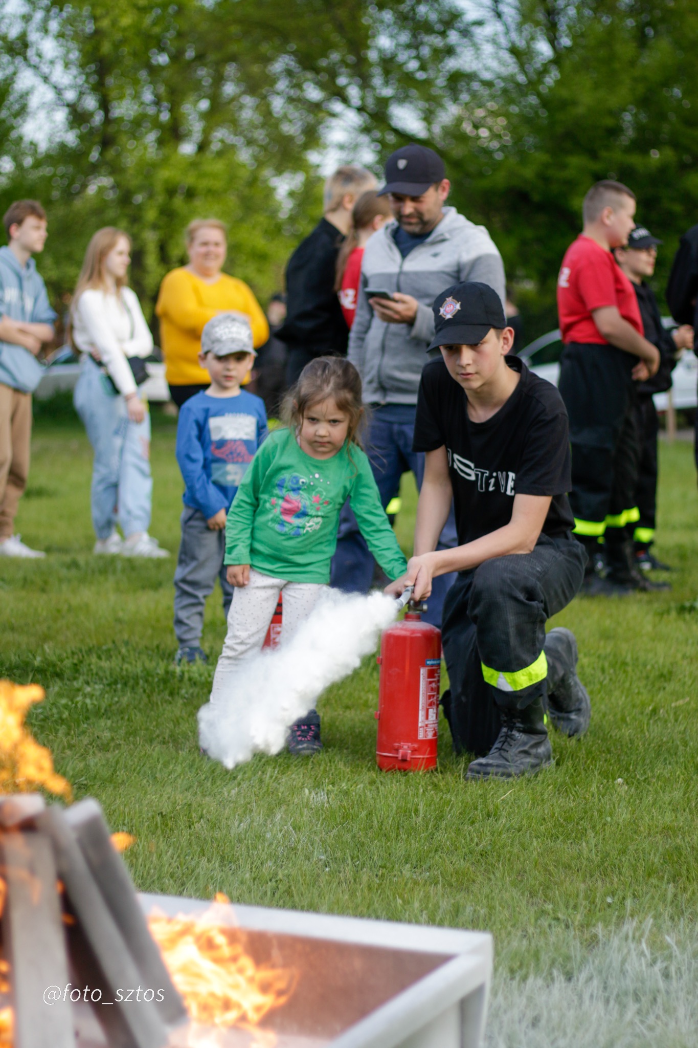 Młody strażak demonstruje użycie gaśnicy proszkowej, celując w płonący metalowy kosz, obok stoi mała dziewczynka w zielonej bluzce, w tle obserwujący.