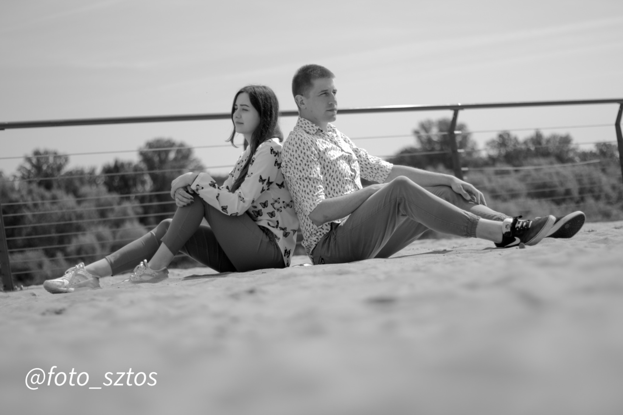 Czarno-białe zdjęcie pary siedzącej plecami do siebie na piasku, z widocznym metalowym ogrodzeniem w tle i podpisem @foto_sztos w lewym dolnym rogu.
