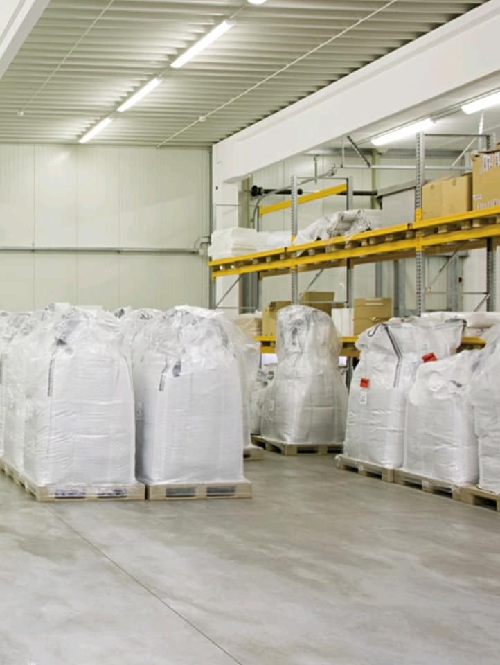 Wnętrze magazynu z rzędami palet z workami, oświetlone lampami jarzeniowymi pod sufitem, widoczne regały magazynowe z kartonami w tle.