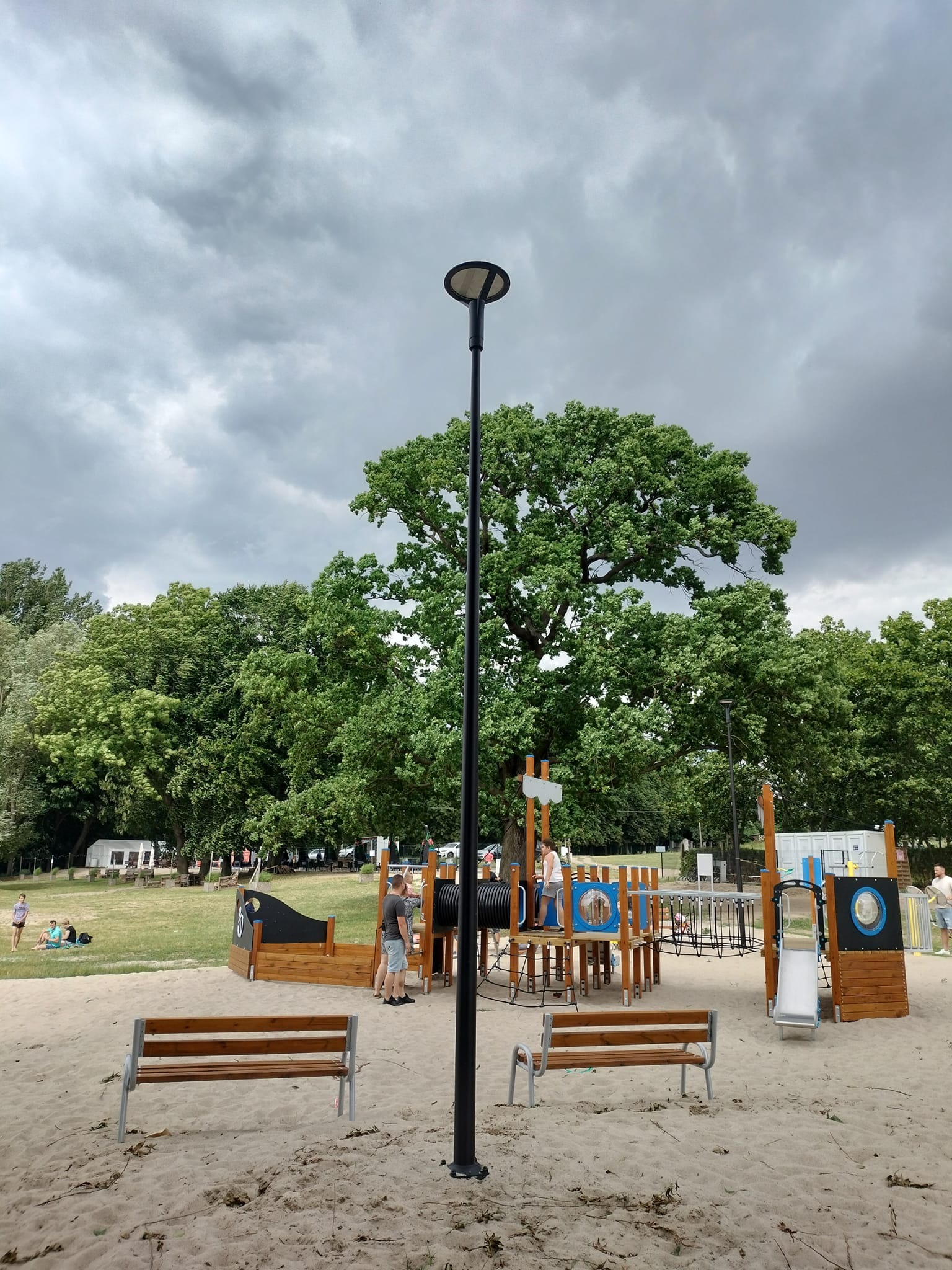 Plac zabaw na piaszczystej powierzchni z drewnianymi konstrukcjami, ławkami i wysoką, czarną latarnią uliczną na tle pochmurnego nieba i zieleni drzew.