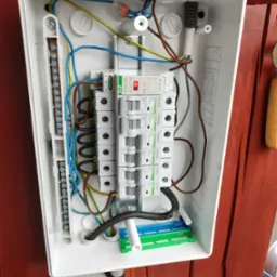 Otwarta skrzynka elektryczna z zamontowanymi bezpiecznikami NOARK, widoczne przewody w różnych kolorach, listwa zaciskowa i złączki, zamocowana na drewnianej ścianie. Czerwona dioda na jednym...