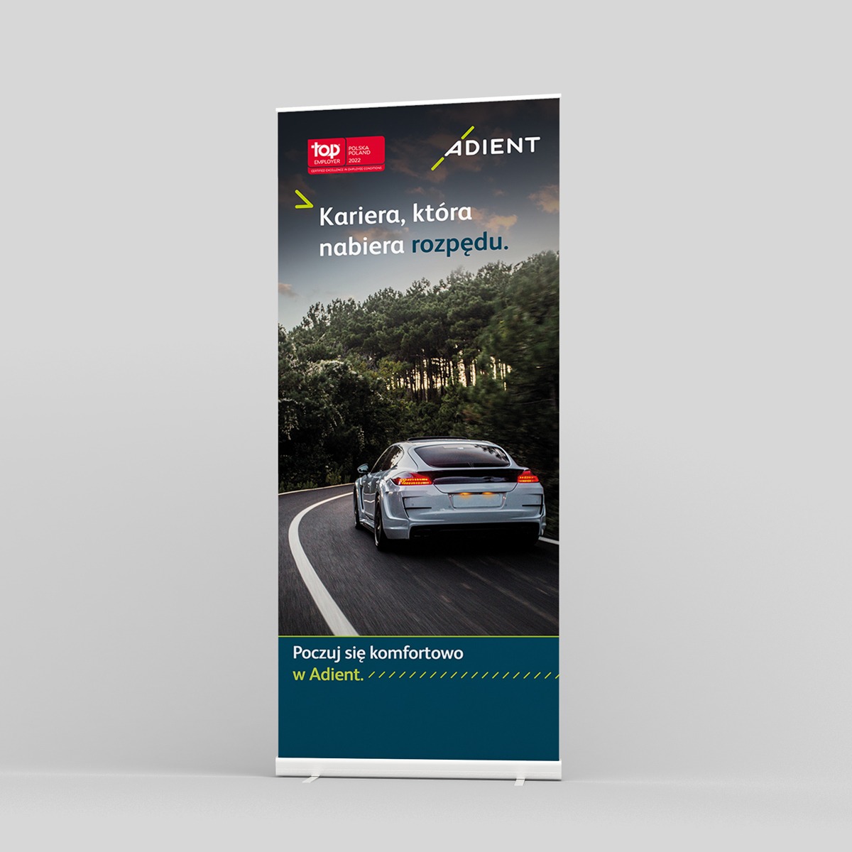 Roll-up reklamowy firmy Adient z hasłem 'Kariera, która nabiera rozpędu' i zdjęciem srebrnego samochodu jadącego krętą drogą.