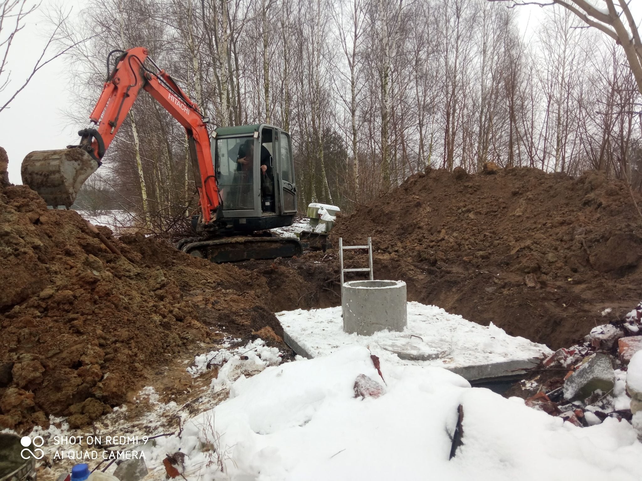Pomarańczowa koparka Hitachi podczas montażu betonowego zbiornika w zimowej scenerii, widoczna drabina i wykopana ziemia.