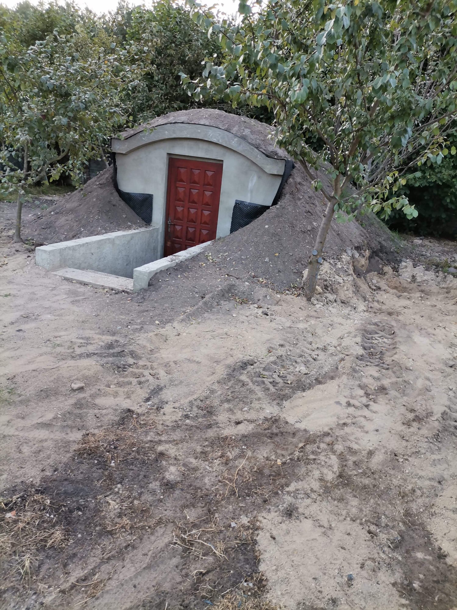 Betonowy schron ziemny z czerwoną, wzorzystą, drewnianą bramą wejściową, częściowo zakopany w ziemi, z betonowymi schodami prowadzącymi do wejścia, widoczne ślady opon na ziemi.