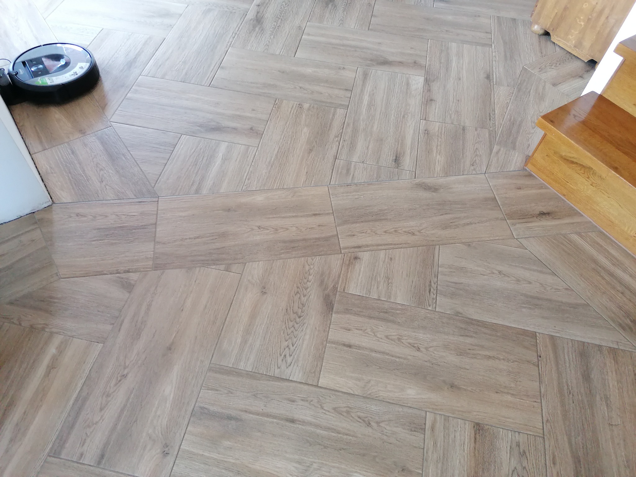 Ułożona w jodełkę podłoga z płytek imitujących drewno, z widocznym robotem sprzątającym i fragmentem drewnianych schodów.