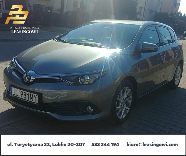Szary samochód osobowy Toyota Auris zaparkowany na chodniku przed budynkiem, z widoczną tablicą rejestracyjną LU 981MY i logotypem firmy leasingowej w rogu.