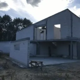 Surowy stan dwukondygnacyjnego budynku w trakcie budowy, wzniesione ściany z pustaków, widoczne otwory okienne i drzwiowe, betonowa wylewka fundamentowa, w tle drzewa i zachmurzone niebo.