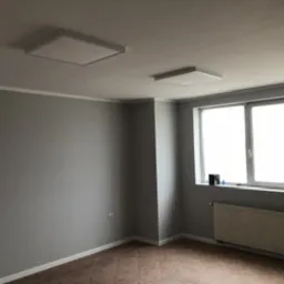 Szare wnętrze pokoju po szpachlowaniu i malowaniu, widoczne gładkie ściany, biały sufit z dwoma kwadratowymi lampami LED, biała listwa przypodłogowa, okno z grzejnikiem i częściowo zakryte parapety.