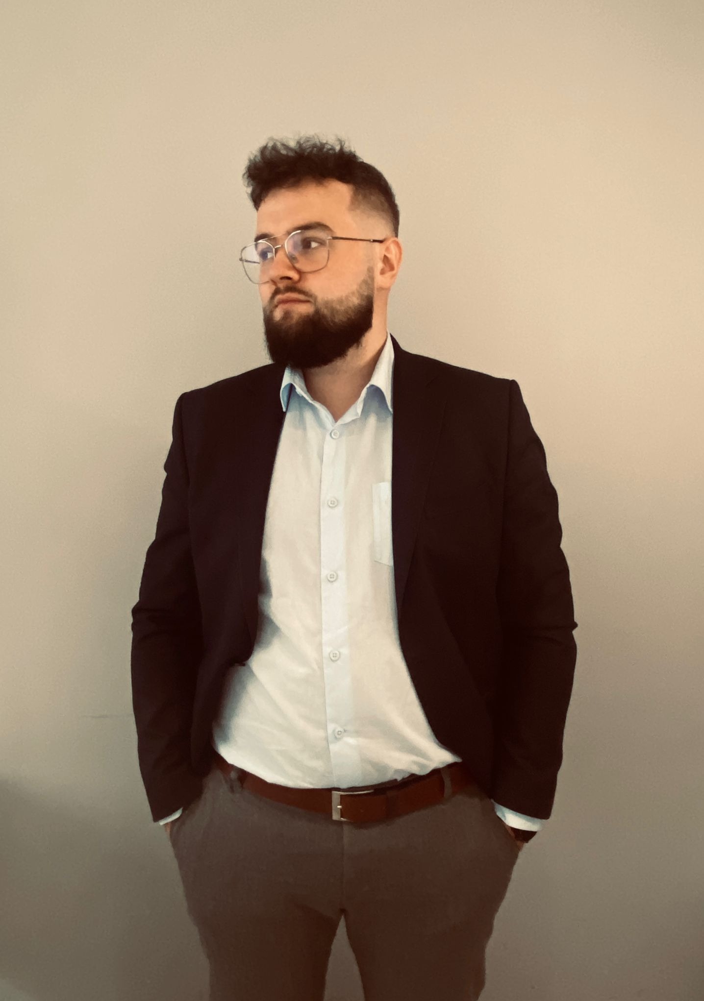 Młody mężczyzna z brodą i okularami, ubrany w białą koszulę i czarną marynarkę, stoi na neutralnym tle, z rękami w kieszeniach spodni w kolorze gołębim, patrząc w lewo.