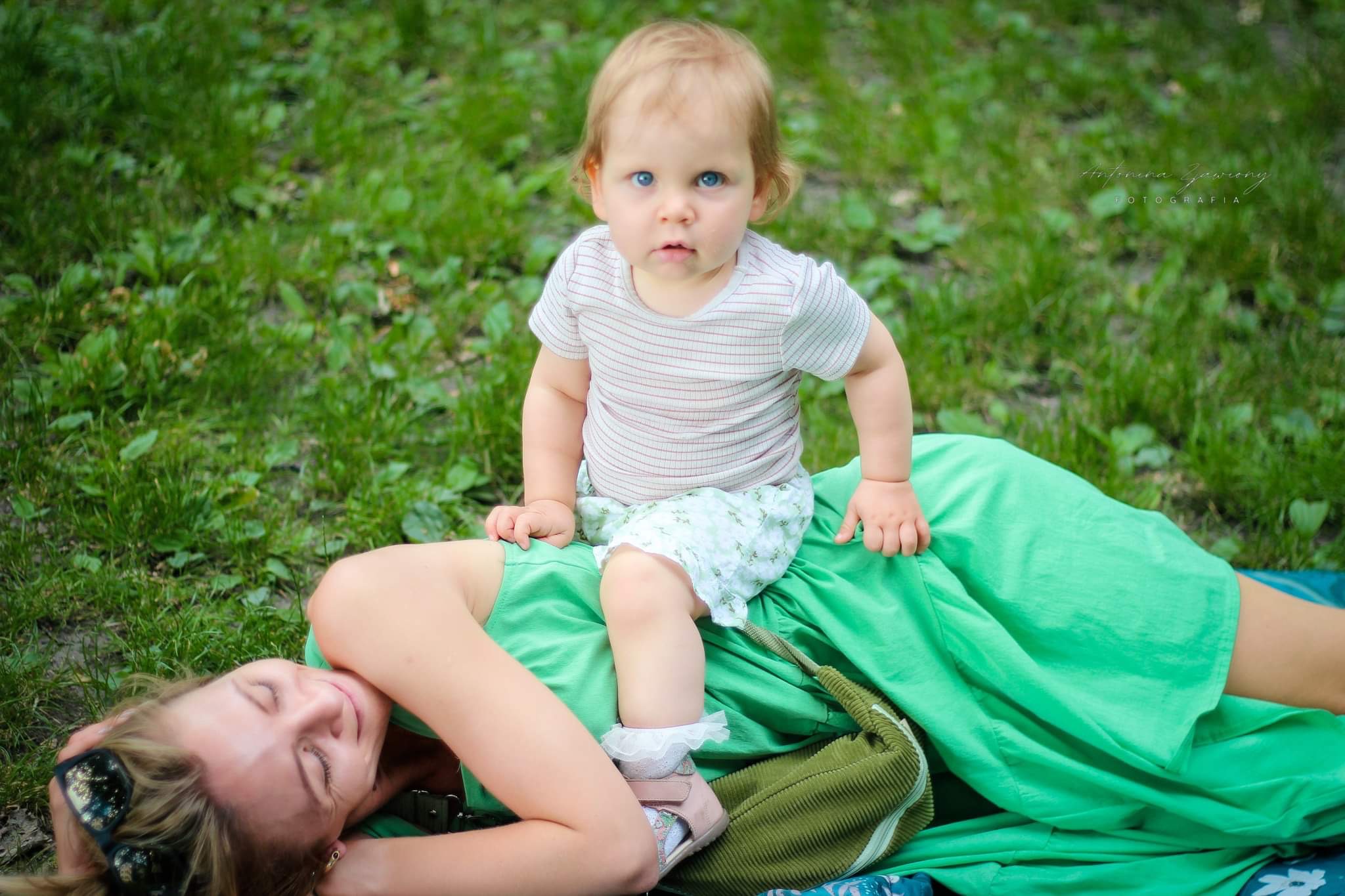 Niemowlę z niebieskimi oczami siedzi na brzuchu uśmiechniętej mamy leżącej na trawie w parku.