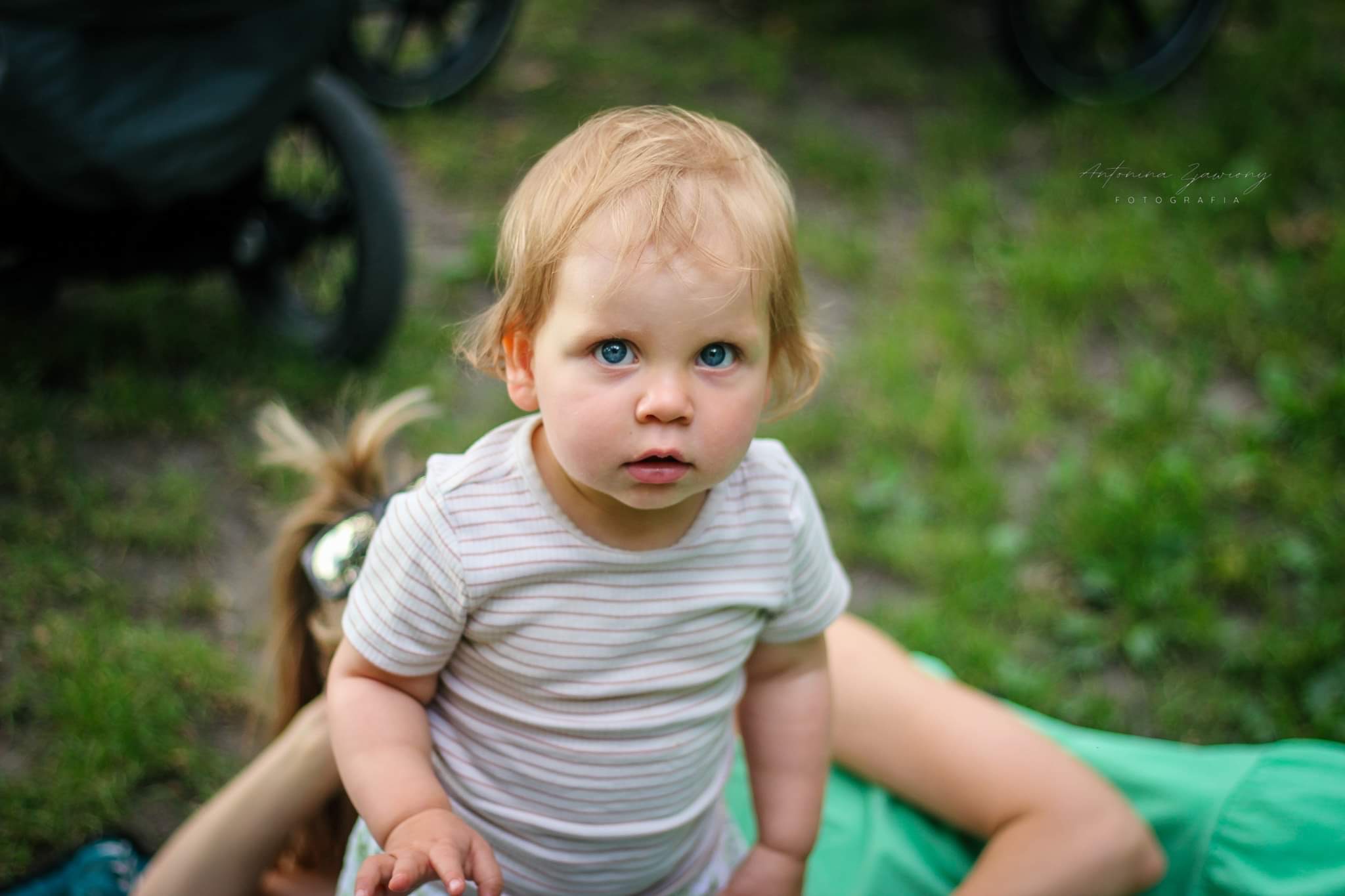 Portret uroczego blondwłosego dziecka z intensywnie niebieskimi oczami, ubranego w koszulkę w paski, patrzącego w górę, z fragmentem wózka dziecięcego i osoby dorosłej w tle na trawie.