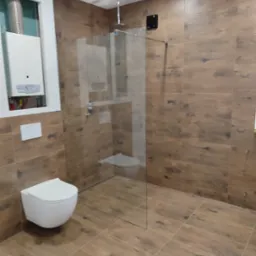 Nowoczesna łazienka z drewnopodobnymi płytkami, kabiną prysznicową typu walk-in, podwieszaną miską WC i piecem gazowym w zabudowie.