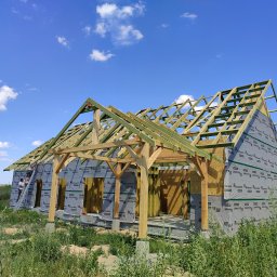 Profibud Krzysztof Siemoński - Budowa domu w technologii szkieletowej: widok na konstrukcję dachu z drewnianych belek, ściany pokryte membraną paroizolacyjną, pracownicy na drabinie oraz bujna roślinność na pierwszym planie.