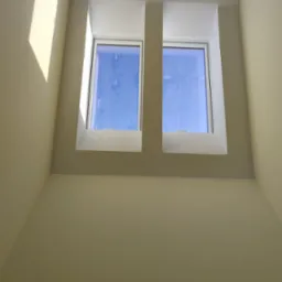 Dwa okna dachowe, widok od wewnątrz, z widocznym niebem i światłem słonecznym padającym na ścianę