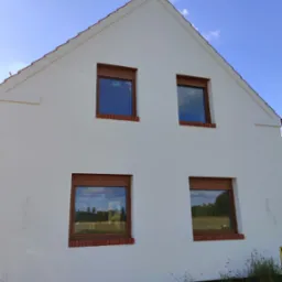 Biała ściana szczytowa domu z czterema oknami z brązowymi ramami i roletami zewnętrznymi, widoczne naklejki na szybach, ceglana podmurówka okien, błękitne niebo w tle.