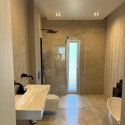 Jan Centkowski - Nowoczesna łazienka z beżowymi płytkami, białym blatem umywalki i czarną armaturą, minimalistyczny design z kabiną prysznicową ze szklaną ścianką i wąskim oknem.