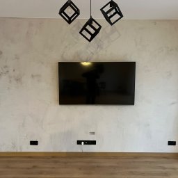 Jan Centkowski - Ściana w salonie z efektem dekoracyjnym, podwieszony telewizor, nowoczesne lampy w kształcie czarnych sześcianów, widoczne gniazdka elektryczne przy podłodze.