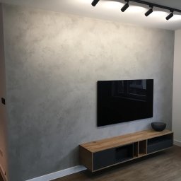 Jan Centkowski - Ściana wykończona dekoracyjną strukturą w odcieniach szarości, oświetlona listwą z reflektorami, z zawieszonym telewizorem i szafką RTV w stylu nowoczesnym.