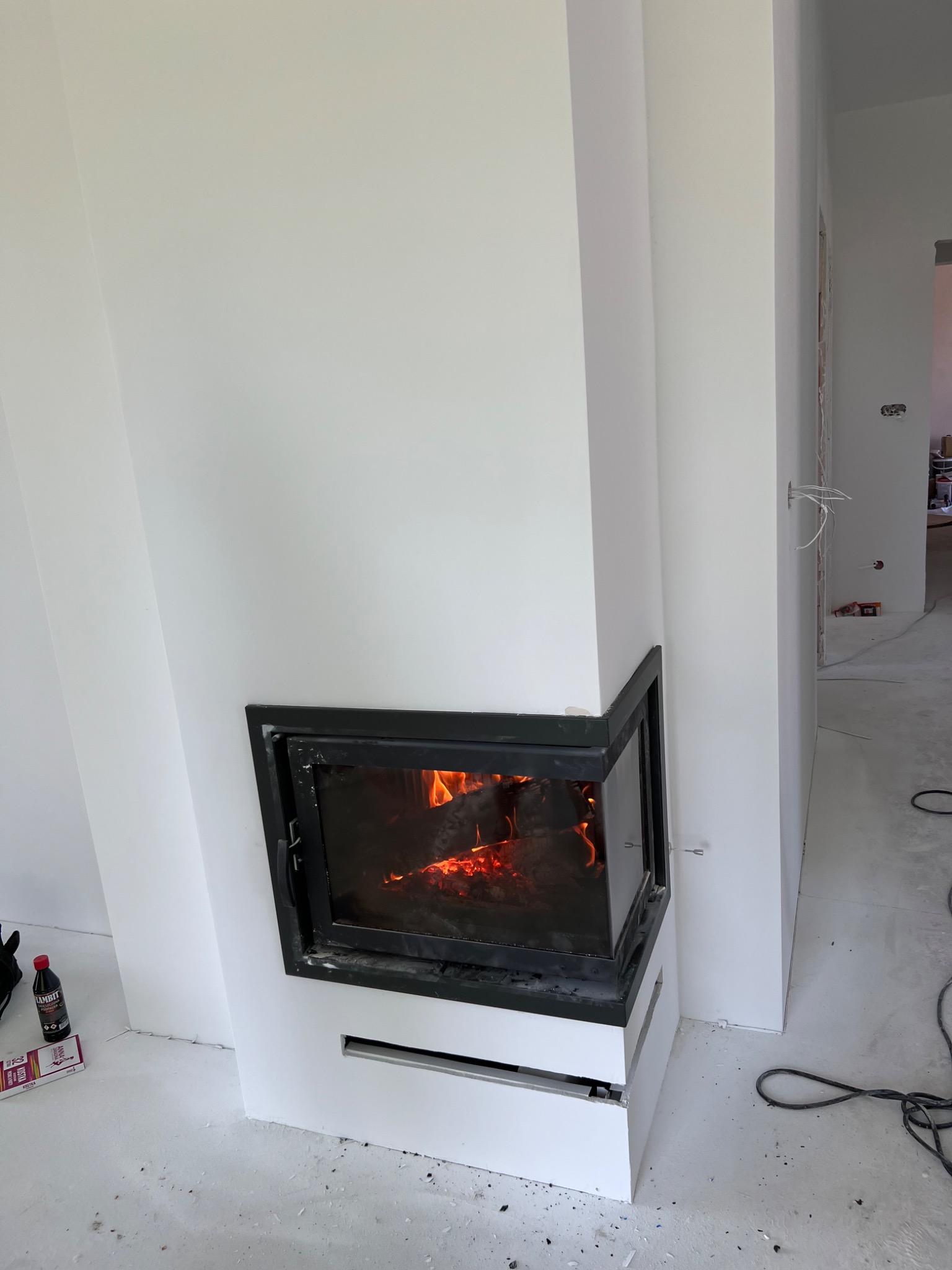 Nowoczesny, biały kominek narożny z czarną ramą i palącym się ogniem, w trakcie wykończenia wnętrza, widoczne niedoskonałości na podłodze i białe ściany.