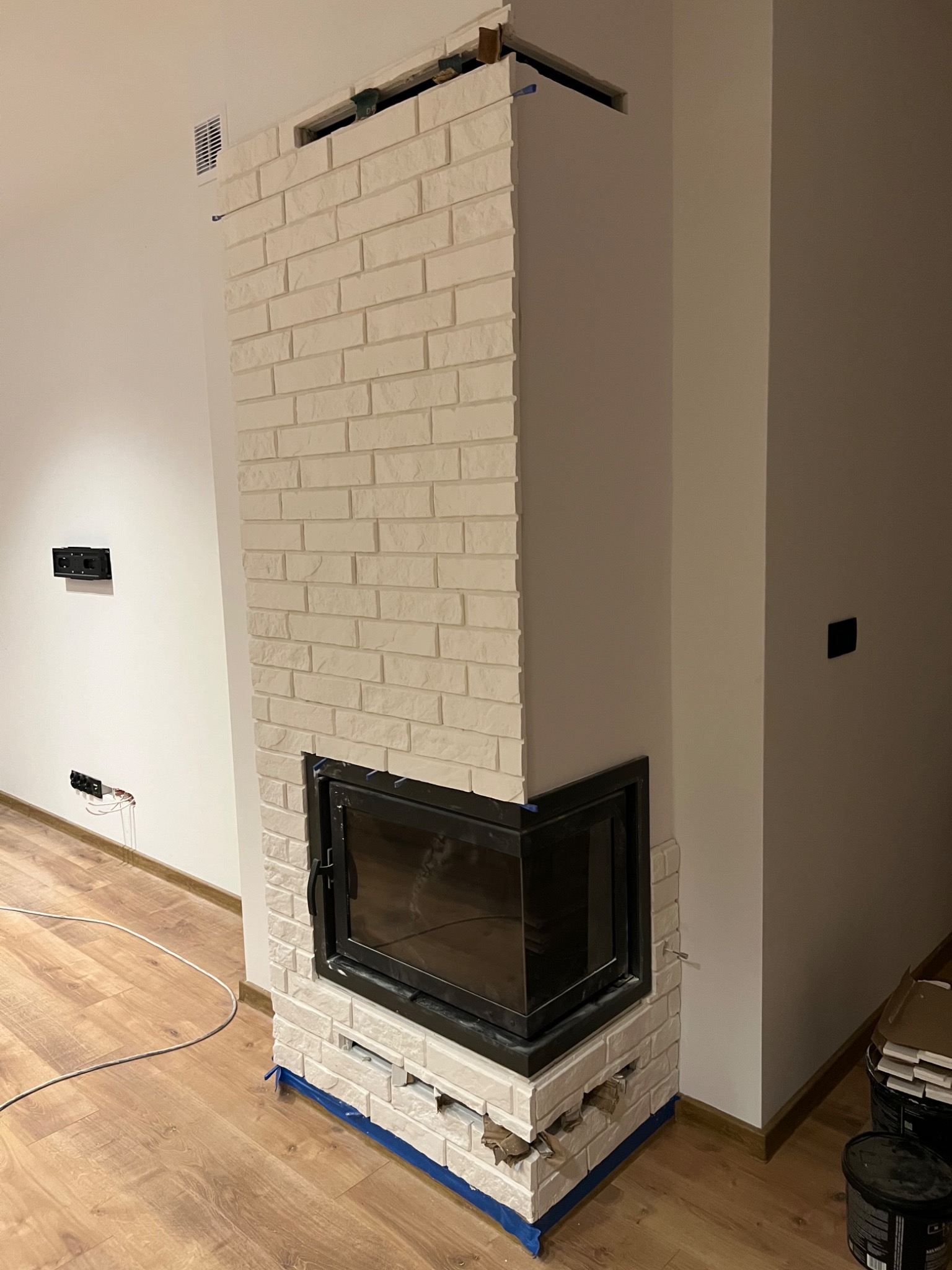 Wnętrze z kominkiem obłożonym białymi cegłami, w trakcie wykończenia, widoczne taśmy malarskie zabezpieczające podłogę i niedokończone fragmenty obudowy.