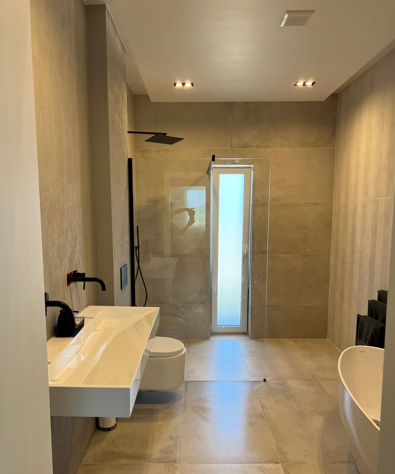 Nowoczesna łazienka z beżowymi płytkami, białym blatem umywalki i czarną armaturą, minimalistyczny design z kabiną prysznicową ze szklaną ścianką i wąskim oknem.