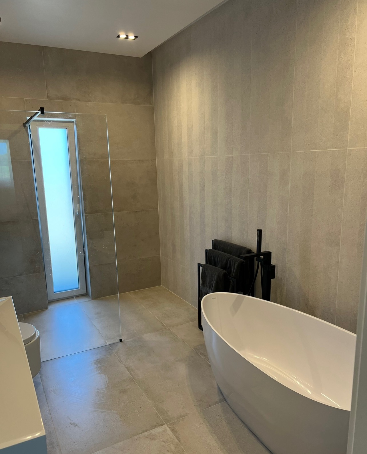 Nowoczesna łazienka z wolnostojącą białą wanną, kabiną prysznicową ze szklaną ścianką i szarą, dużą płytką na ścianach i podłodze.